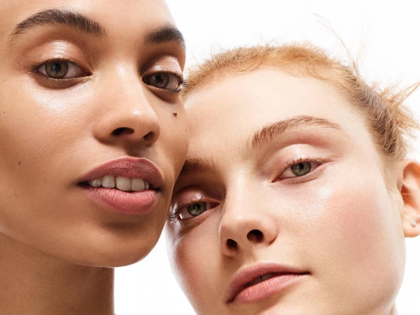 Close-up studio portrait of two women with fair skin tones, soft lighting, and subtle makeup against a stark white backdrop.