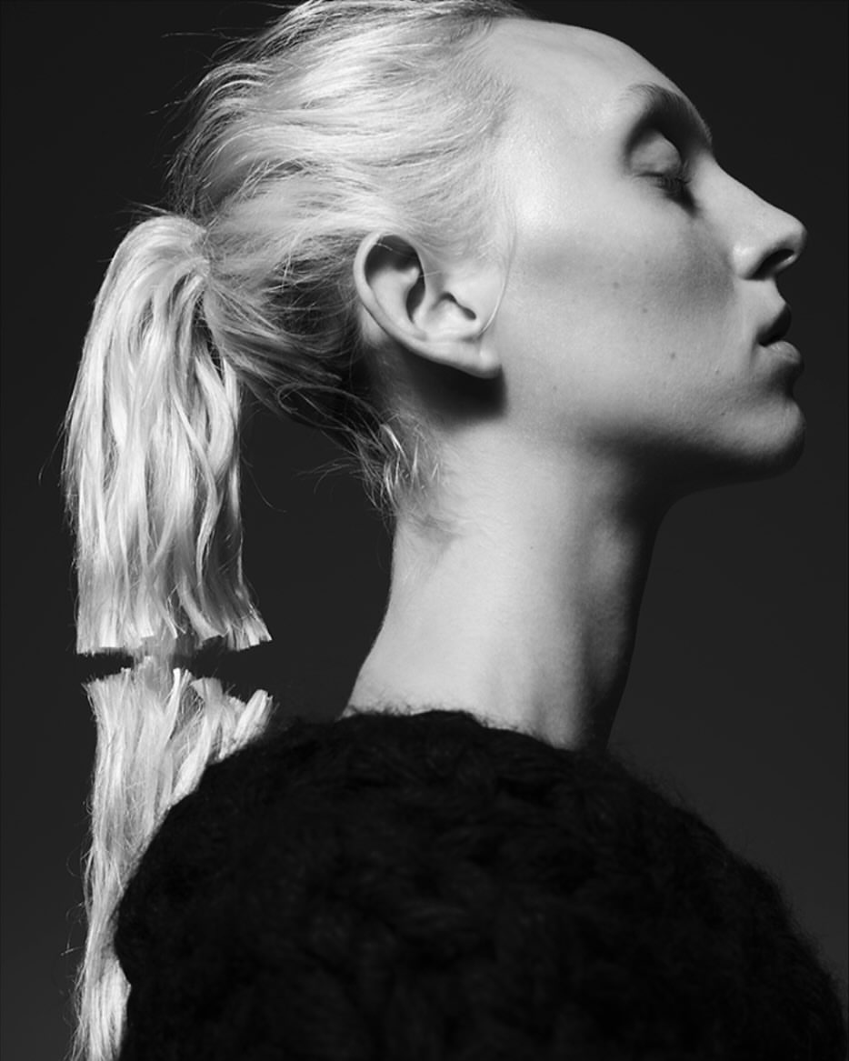 Black and white profile portrait of a woman with blonde hair in a low ponytail and a dark textured sweater against a dark background.