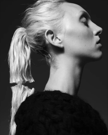 Black and white profile portrait of a woman with blonde hair in a low ponytail and a dark textured sweater against a dark background.