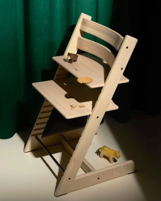 Light wood high chair with snacks and a toy animal on the floor in front of a green and black background, product photography.