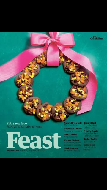 Chocolate cookies with dried fruit and nuts are arranged in a wreath formation, adorned with a pink ribbon on a teal background.