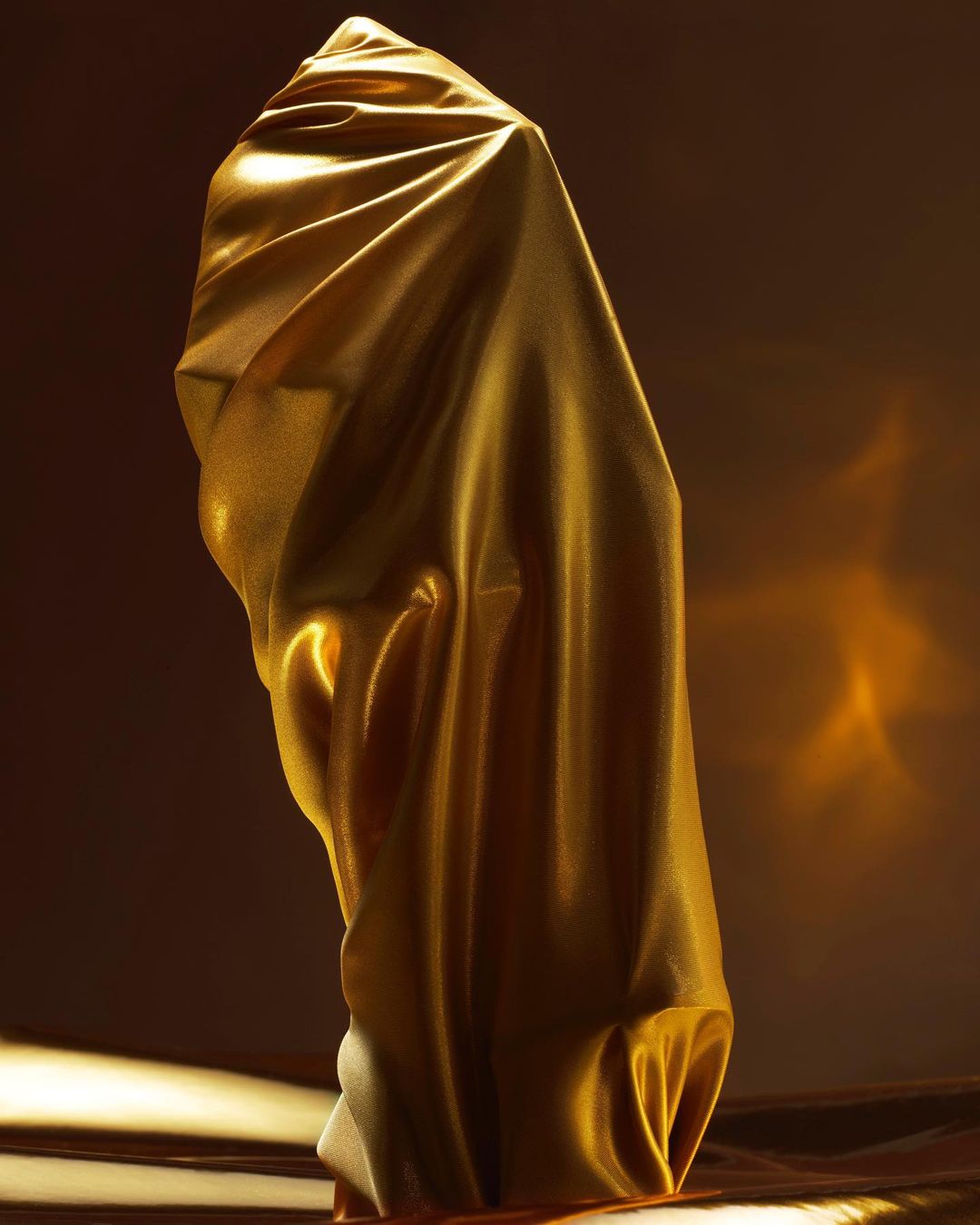 Abstract digital rendering: Gold-colored fabric drapes over a form, with soft light creating depth and texture against a dark background.