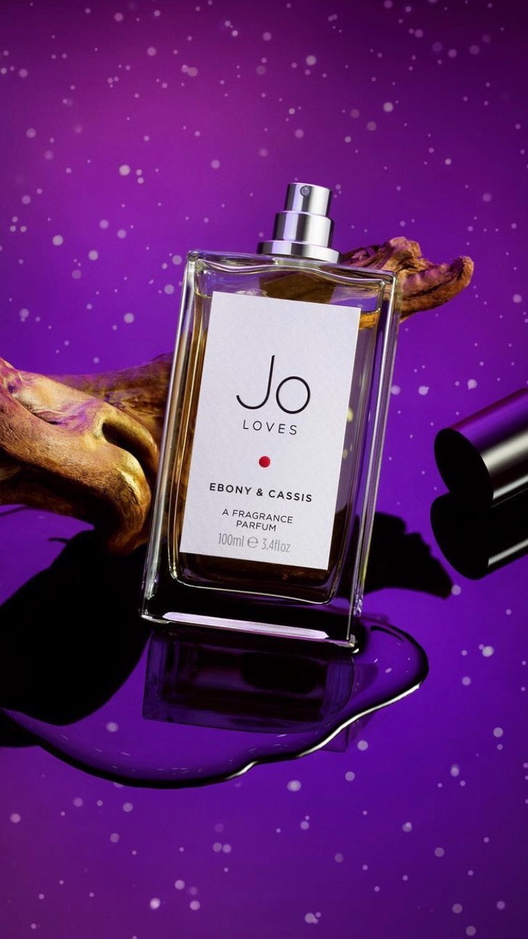 Product shot of Jo Loves Ebony & Cassis perfume bottle against purple background with artistic gold driftwood and liquid spill.