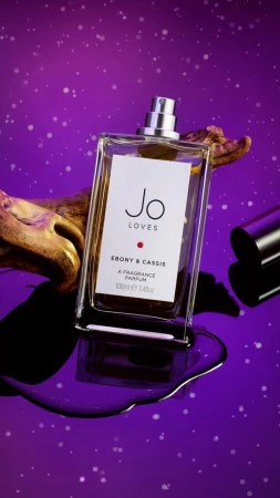 Product shot of Jo Loves Ebony & Cassis perfume bottle against purple background with artistic gold driftwood and liquid spill.
