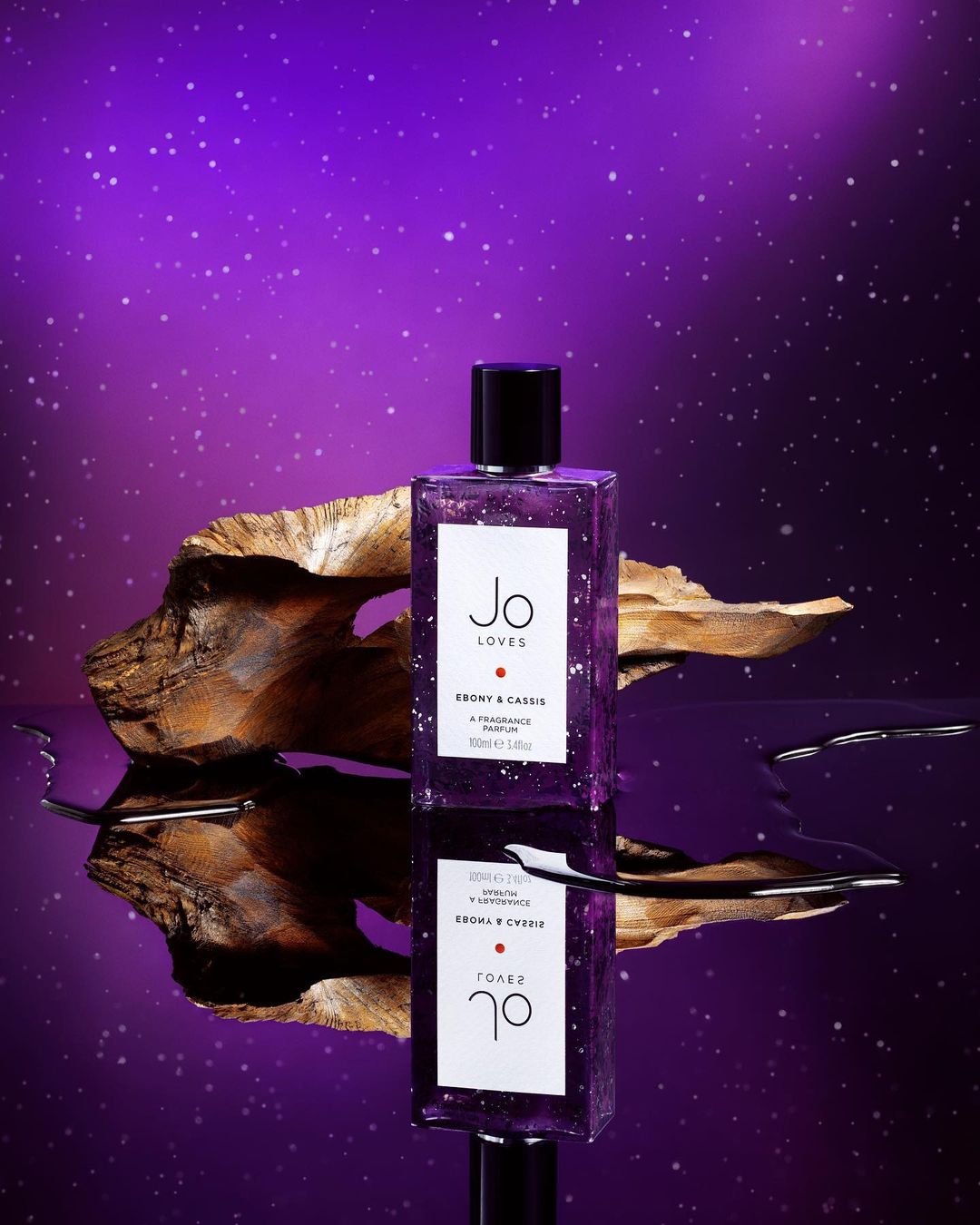 A purple-tinted perfume bottle by Jo Loves is reflected on a surface, next to a piece of wood, set against a gradient purple background.