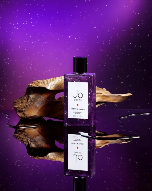 A purple-tinted perfume bottle by Jo Loves is reflected on a surface, next to a piece of wood, set against a gradient purple background.