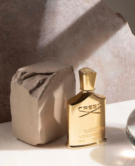 Close-up of a gold Creed fragrance bottle against a textured beige backdrop with soft lighting, highlighting luxury and sophistication.