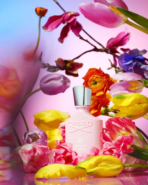 Still life shot of a pink Creed perfume bottle surrounded by colorful tulips and other flowers against a gradient pink and blue background.