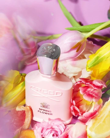 Creed Spring Flower perfume bottle on a purple background surrounded by pink, yellow and white tulips and citrus slices.