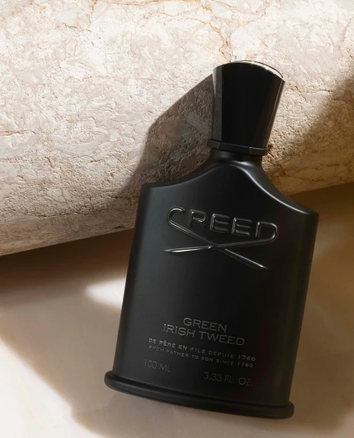 Creed Green Irish Tweed black perfume bottle on light surface with white rock behind, close-up product shot in beauty advertisement.