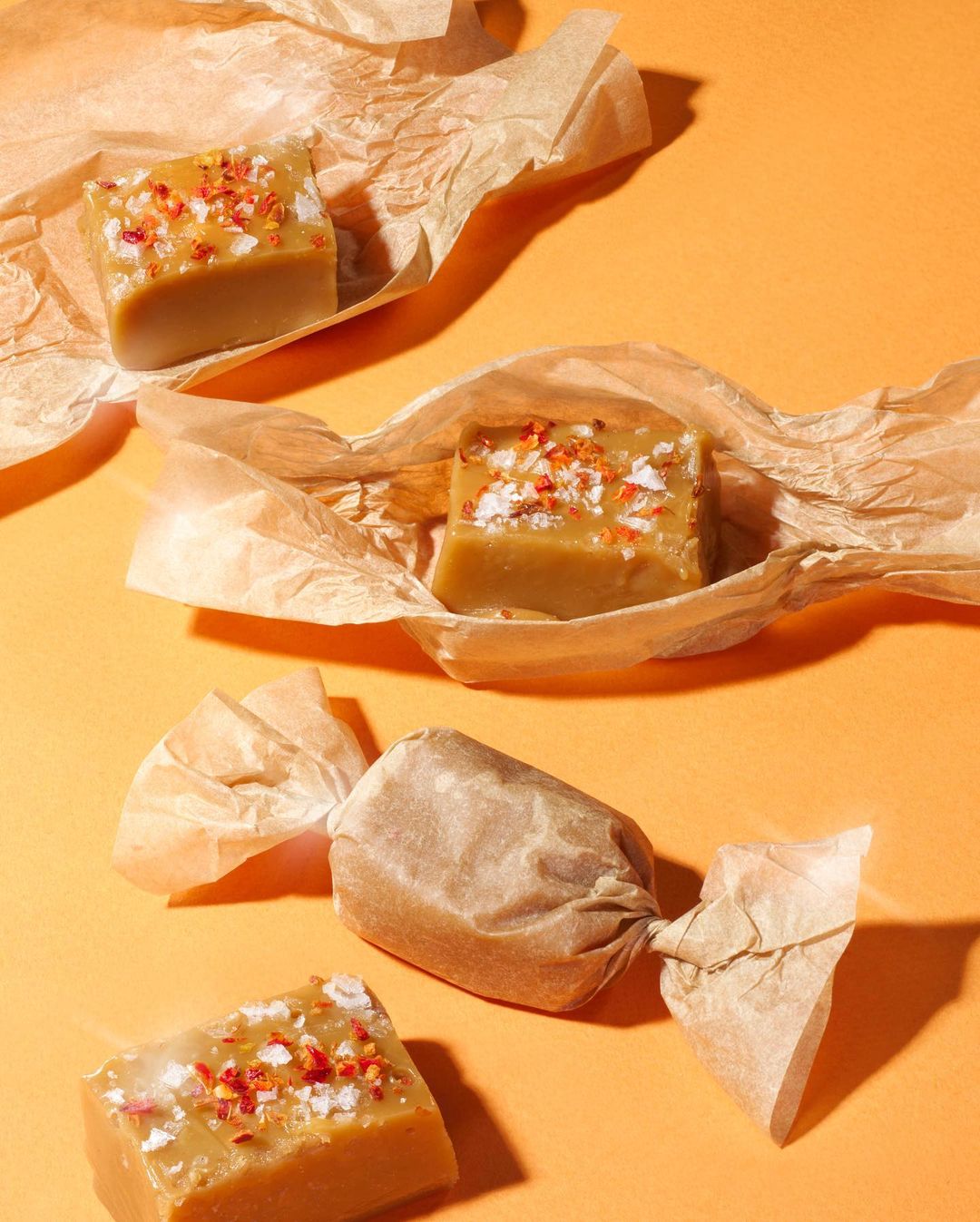 Studio shot of four caramel candies wrapped in crumpled wax paper on an orange background, showcasing the texture and color of the treats.