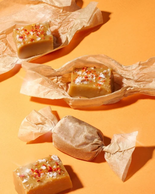Studio shot of four caramel candies wrapped in crumpled wax paper on an orange background, showcasing the texture and color of the treats.