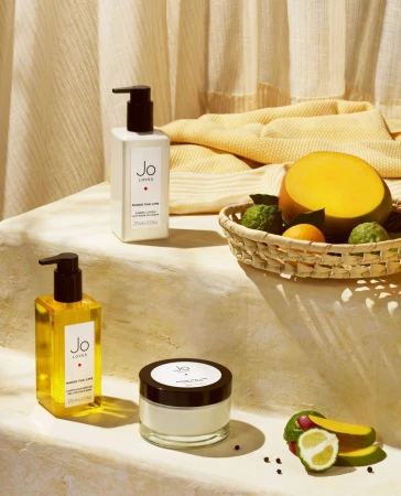 Still life featuring Jo Loves bath and body products arranged with fresh fruit, including mango and citrus, in a naturally lit studio setting.