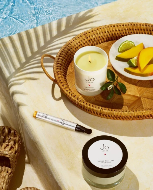 Jo Loves candle, perfume, and body cream are staged on a woven tray with mango and lime near a pool, under palm frond shadows.