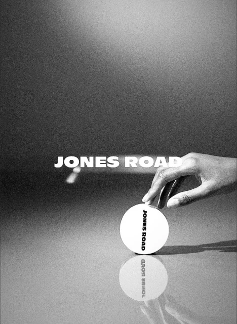 Monochrome product shot of Jones Road beauty product with dark-skinned hand, on reflective surface, against gray gradient backdrop.