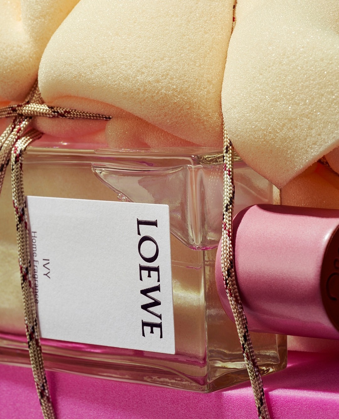Loewe Ivy home fragrance displayed in a glass bottle with a pink cap and decorative rope, set against a pink background and beige foam.