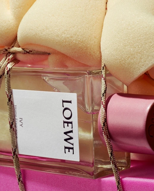 Loewe Ivy home fragrance displayed in a glass bottle with a pink cap and decorative rope, set against a pink background and beige foam.