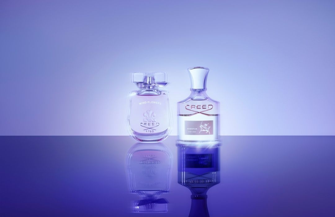 Two Creed perfume bottles, one Wind Flowers and one Aventus, stand on a reflective surface against a graduated purple backdrop in a studio shot.
