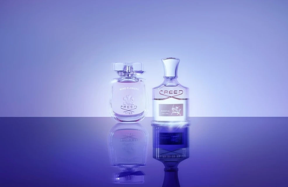Two Creed perfume bottles, one Wind Flowers and one Aventus, stand on a reflective surface against a graduated purple backdrop in a studio shot.