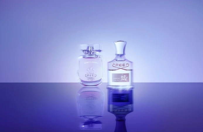 Two Creed perfume bottles, one Wind Flowers and one Aventus, stand on a reflective surface against a graduated purple backdrop in a studio shot.