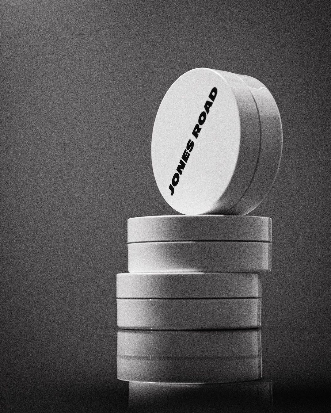 A stack of white cosmetic containers with the brand name JONES ROAD printed on the top, displayed against a monochromatic background.