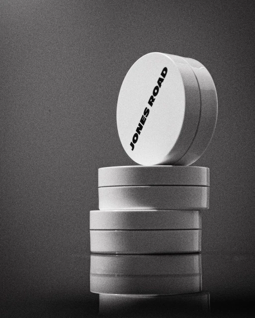 A stack of white cosmetic containers with the brand name JONES ROAD printed on the top, displayed against a monochromatic background.