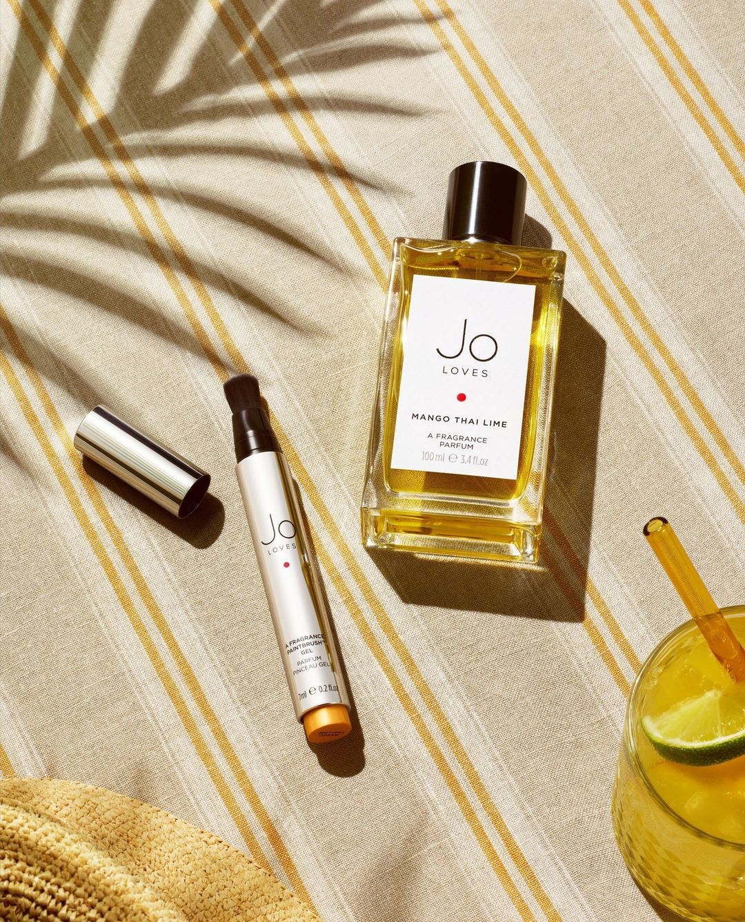 Perfume bottles and a cocktail glass rest on a striped surface in a flat lay with natural light casting shadows, creating a summer vibe.