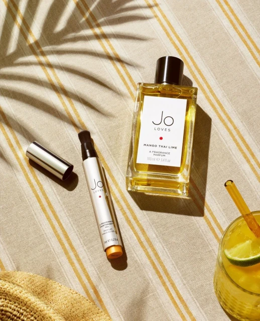 Perfume bottles and a cocktail glass rest on a striped surface in a flat lay with natural light casting shadows, creating a summer vibe.