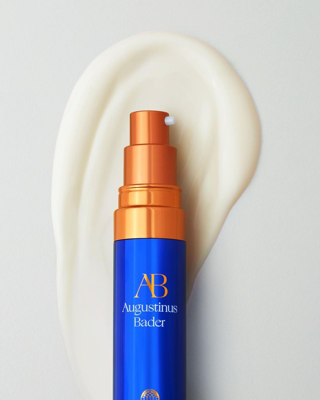 Close-up of a blue and gold Augustinus Bader bottle on white surface with smear of lotion, studio lighting, beauty product shot.