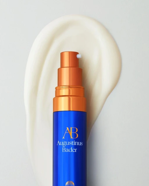 Close-up of a blue and gold Augustinus Bader bottle on white surface with smear of lotion, studio lighting, beauty product shot.