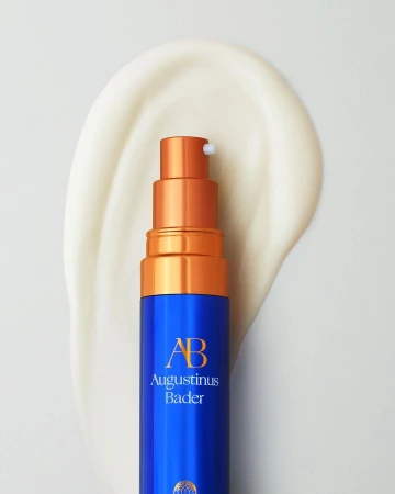 Close-up of a blue and gold Augustinus Bader bottle on white surface with smear of lotion, studio lighting, beauty product shot.
