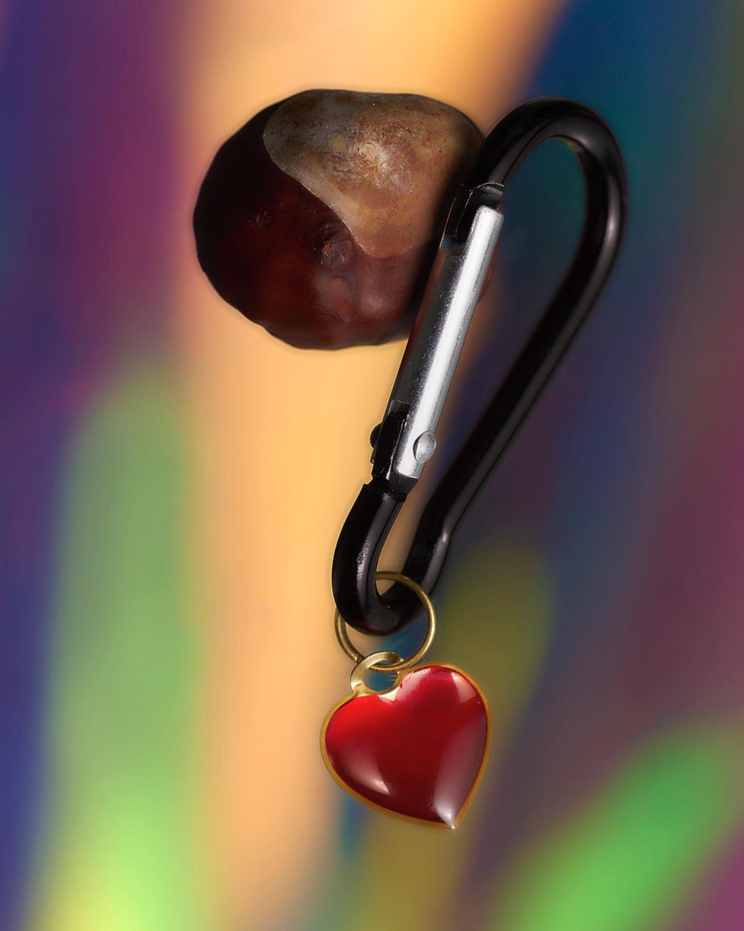 Close-up of a carabiner with a red heart pendant and a chestnut attached, set against a blurry, vibrant, multicolored background.
