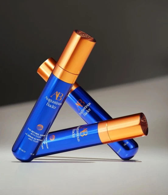Three blue and copper Augustinus Bader The Retinol Serum bottles in a triangular arrangement against a blurred gradient background.