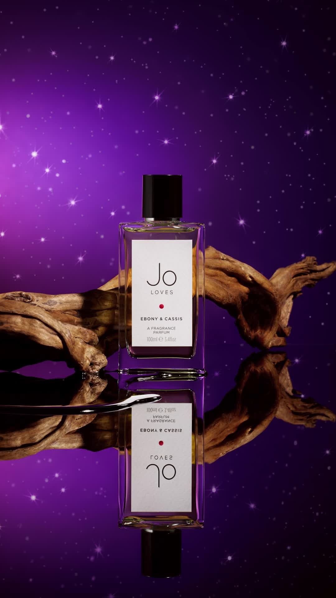 Jo Loves fragrance bottle on a reflective surface with a piece of driftwood in a studio setting with a purple and white backdrop.