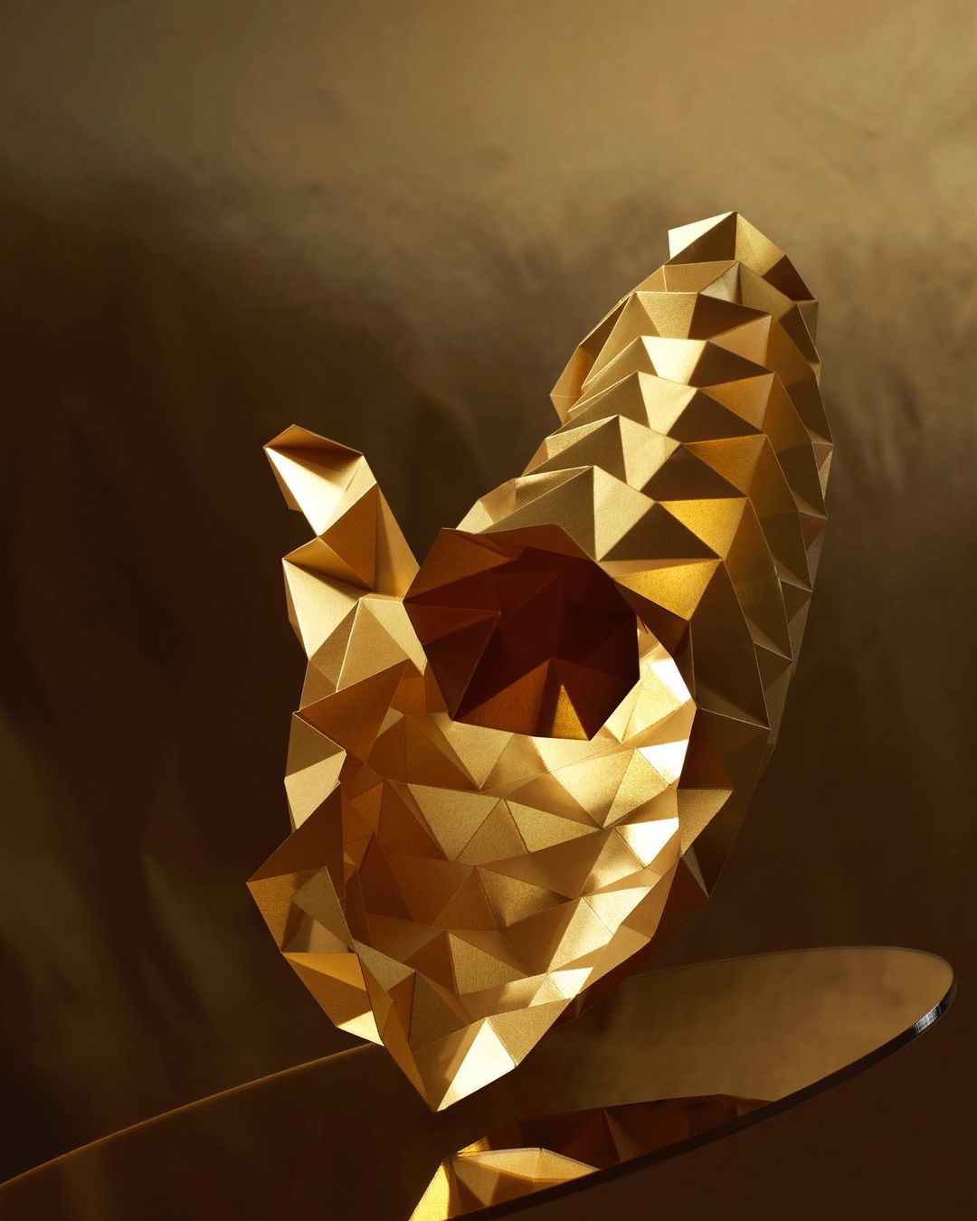 Abstract gold sculpture with geometric, triangular facets against a hazy gold background, creating a shimmering, dimensional effect.