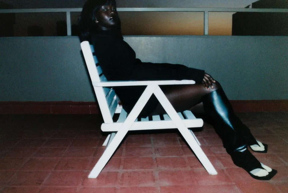 Elegant woman sits on a white chair outside on a tiled floor at night, wearing black top and leggings, with a lit-up window in the background.