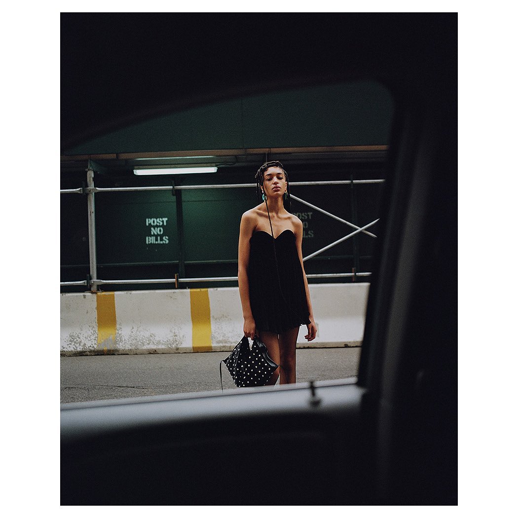 Woman in black strapless dress and carrying a polka dot purse, standing on the street in front of metal guardrail.