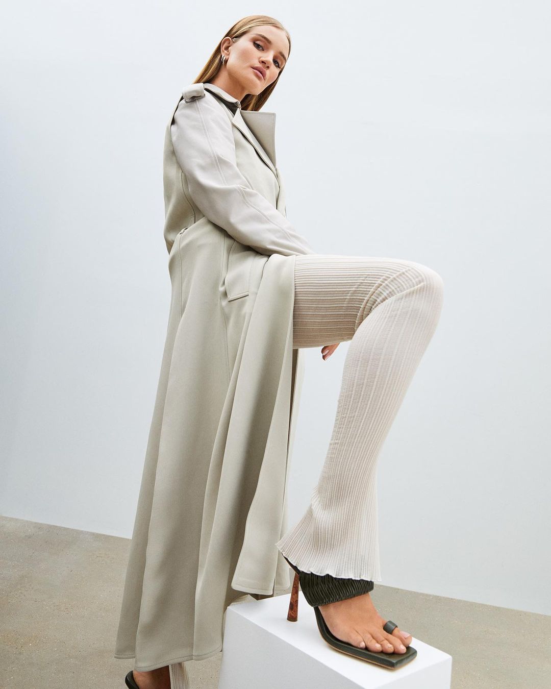 A woman poses in a long beige coat, textured pants, and heels while standing on a white cube in front of a white background.