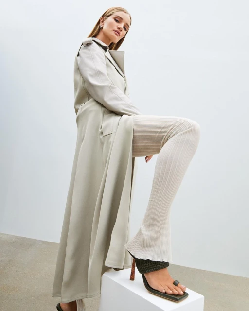 A woman poses in a long beige coat, textured pants, and heels while standing on a white cube in front of a white background.