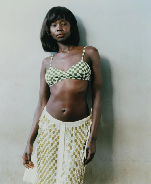Woman in checkered bikini top and fringed skirt stands indoors against neutral backdrop in medium shot with soft lighting and relaxed pose.