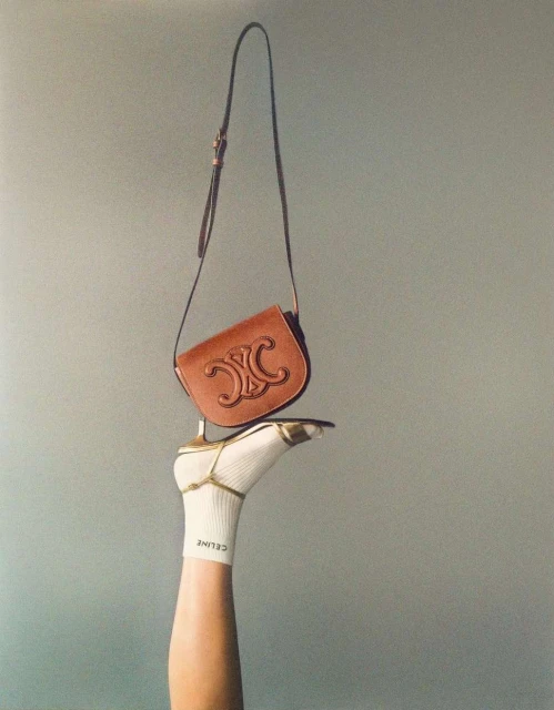 Celine handbag displayed on the sole of a sock-covered foot wearing a gold high-heeled sandal against a gradient gray background.