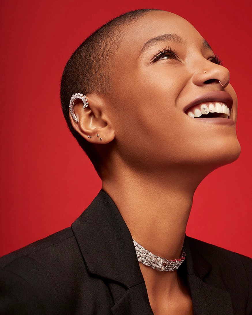 Portrait of a smiling dark-skinned woman with a shaved head, ear cuff, and necklace, posing in front of a red background. Beauty shot.