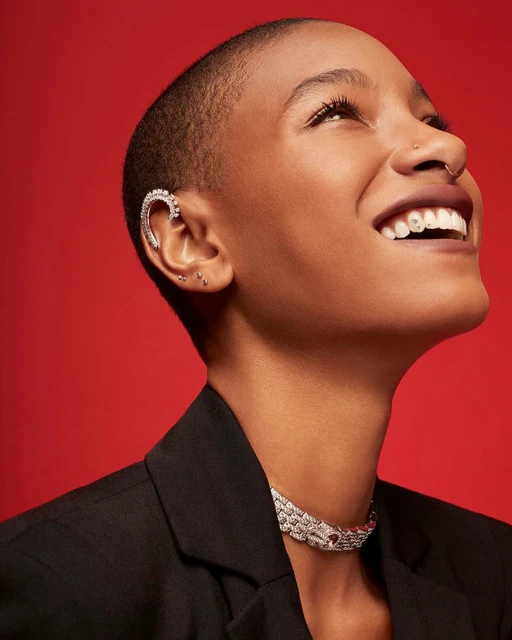Portrait of a smiling dark-skinned woman with a shaved head, ear cuff, and necklace, posing in front of a red background. Beauty shot.