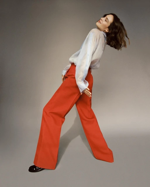 A woman with short brown hair strikes a dramatic pose in a studio wearing a sheer blue blouse and bright red wide-legged pants.