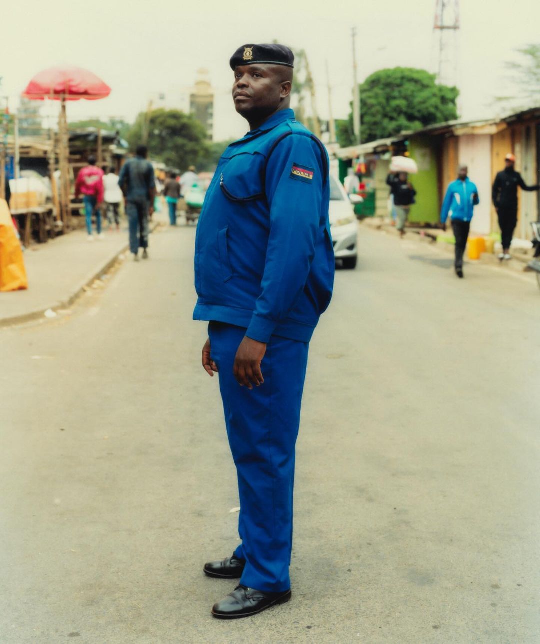 A uniformed guard stands on a street, in medium shot, near an informal market with people walking. The background shows urban buildings.