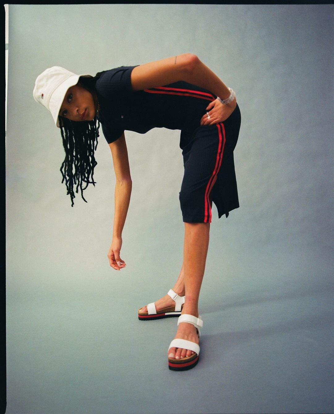 Young woman in a black T-shirt and matching shorts with red stripes, white sandals, and a bucket hat poses indoors.