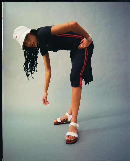 Young woman in a black T-shirt and matching shorts with red stripes, white sandals, and a bucket hat poses indoors.