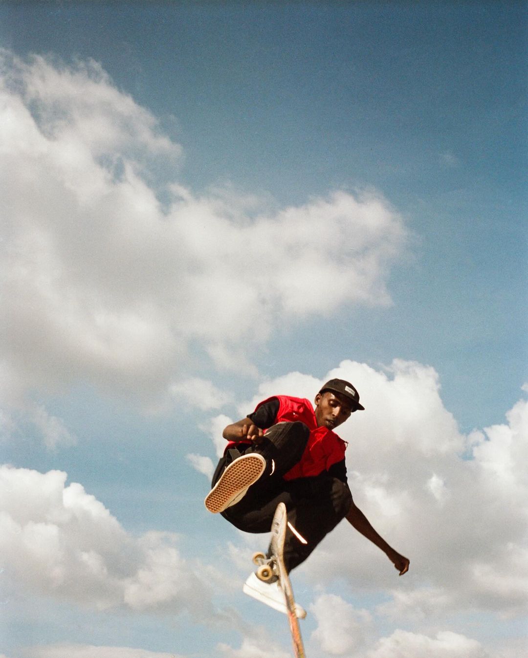 A person performs a skateboard trick in mid-air wearing casual clothes, with a bright blue sky and white clouds in the background.