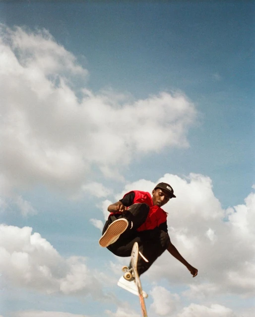 A person performs a skateboard trick in mid-air wearing casual clothes, with a bright blue sky and white clouds in the background.