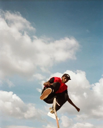 A person performs a skateboard trick in mid-air wearing casual clothes, with a bright blue sky and white clouds in the background.